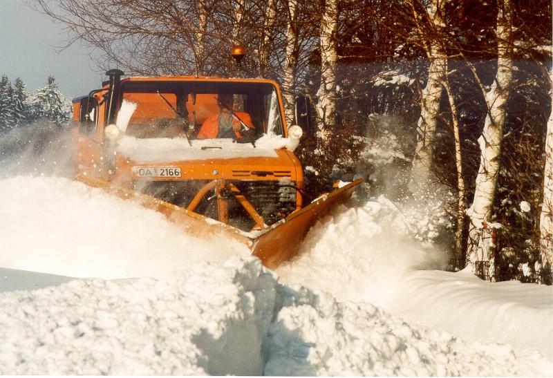 Ein alter orange-farbener Unimog fährt mit einem Spitzpflug durch hohen Schnee