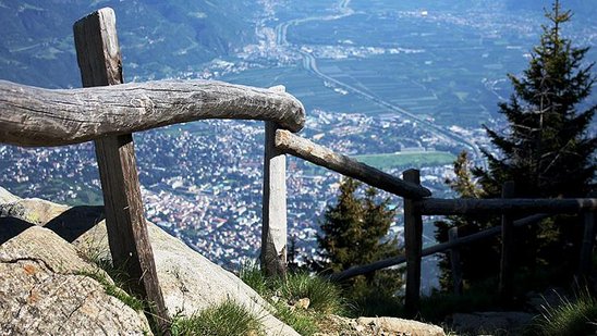 Bergwanderweg mit Holzgeländer mit Blick auf ein besiedeltes Tal.