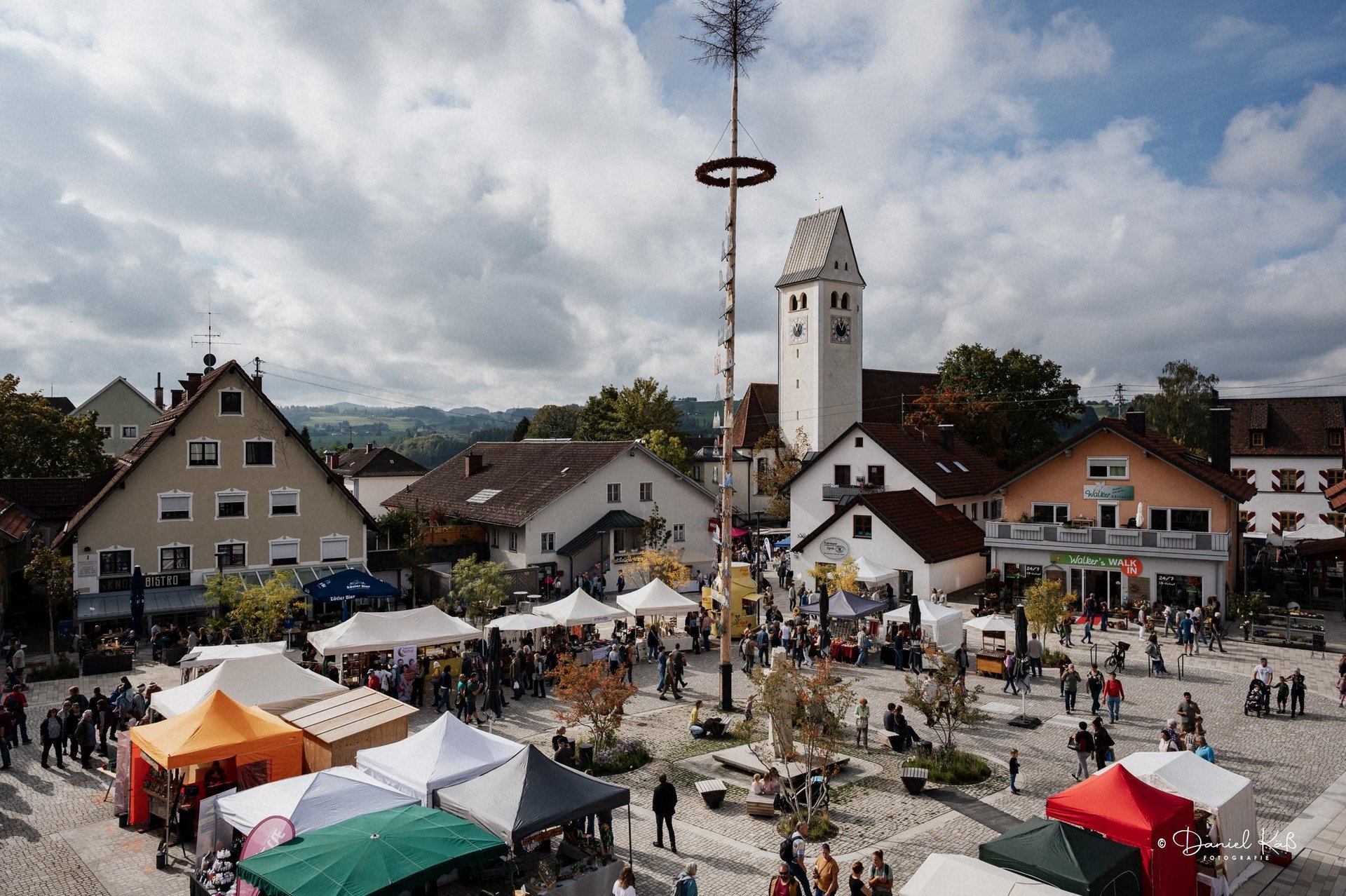 Buntes Markttreiben auf dem Alternativen Markt in Altusried: Zwischen zahlreichen Verkaufsständen tummeln sich Besucherinnen und Besucher auf dem neu gestalteten Marktplatz. Die Pfarrkirche St. Blasius und die Allgäuer Landschaft bilden die stimmungsvolle Kulisse.